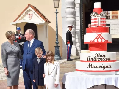 Prince Albert staring at Princess Charlene with their children beside them and a giant birthday cake at the Palace Square