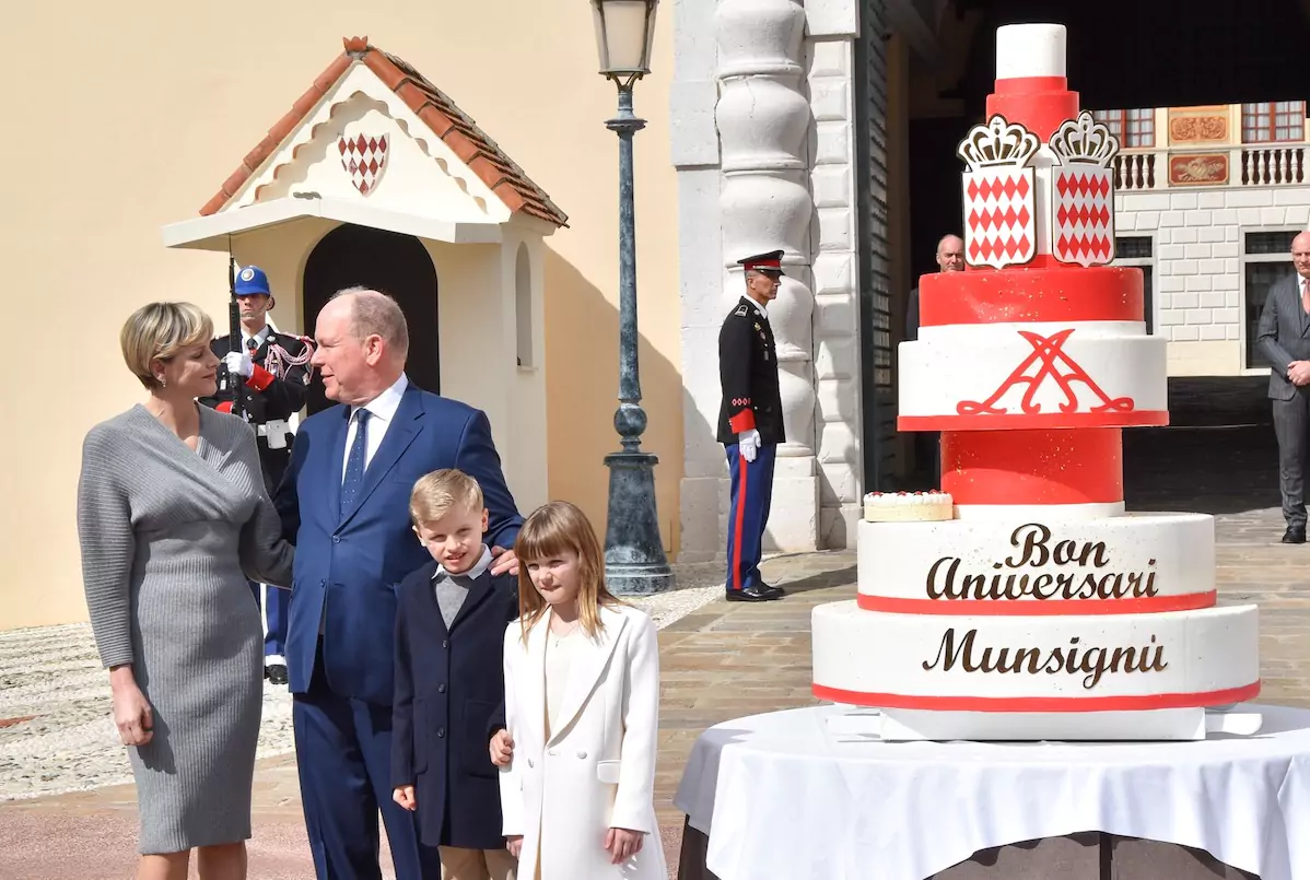 Prince Albert staring at Princess Charlene with their children beside them and a giant birthday cake at the Palace Square